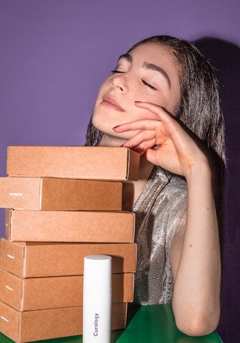 A Gen Z influencer next to stacked boxes of product in front of a purple background, by Curology.