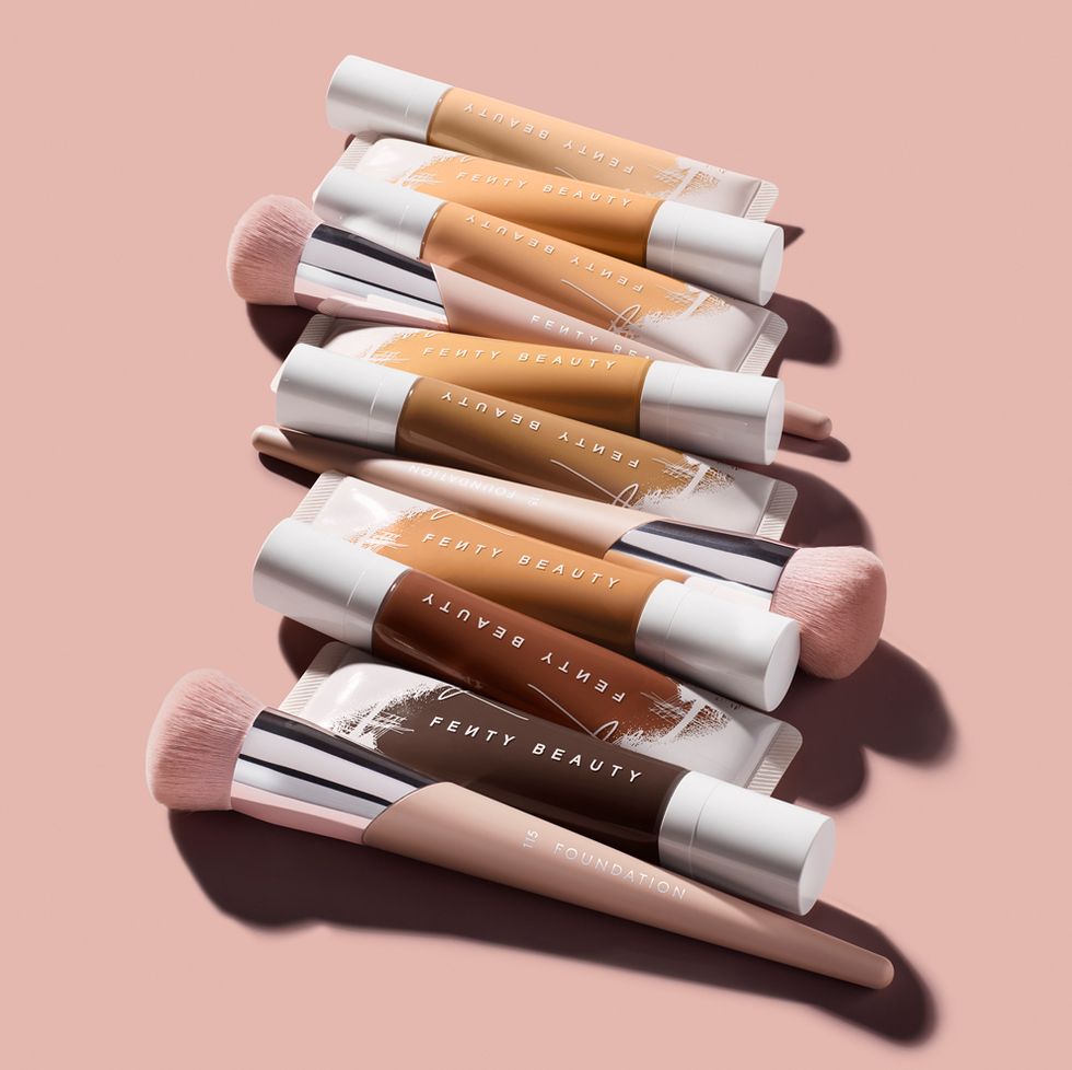 Bottles of Fenty Beauty’s Pro Filt’r Hydrating Longwear Foundation stacked with makeup brushes against a pink background. 