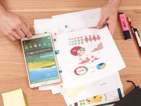 A tablet and several papers with marketing charts arranged on a desk with pens, by Firmbee.com via Unsplash.