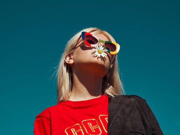 A TikTok influencer wearing colorful glasses with a daisy in her mouth, by Katsiaryna Endruszkiewicz via Unsplash.