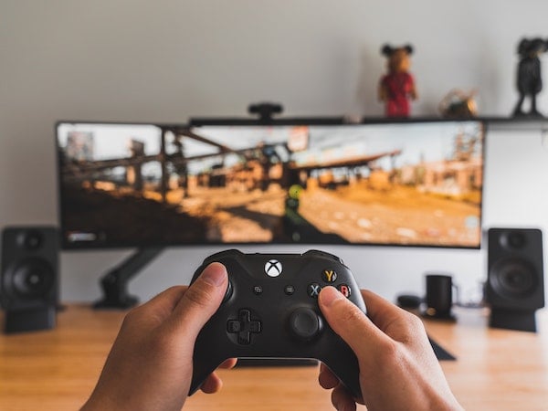 The hands of a gaming creator holding a video game controller in front of a tv screen displaying a shooting game, by Sam Pak via Unsplash.