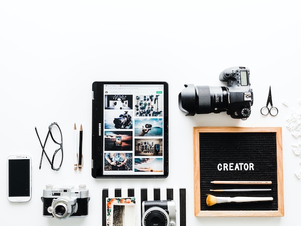 Various objects related to TikTok affiliate marketing, including an iPad, Smartphone, cameras, and a decorative board with the word "creator" in block letters, arranged against a white background, by Brooke Lark via Unsplash.