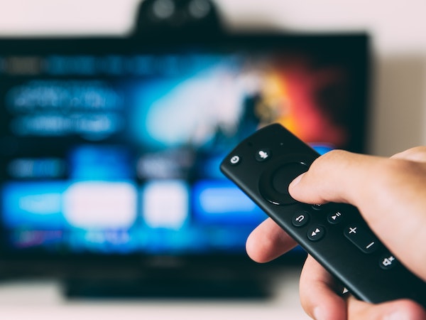 A hand holding a TV remote in front of a TV displaying a show from a media company that is a leader in influencer marketing strategy, by Glenn Carstens Peters via Unsplash.