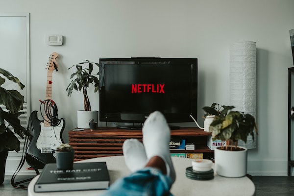 A TV displaying Netflix next to a guitar in a young person's living room, by Mollie Siveram via Unsplash.