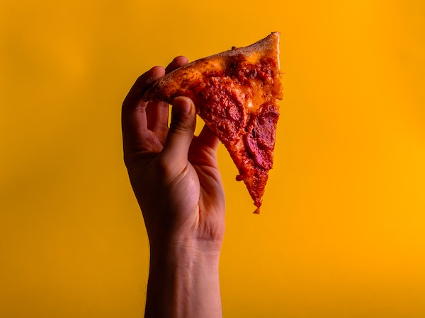 A person's hand holding a DiGiorno pizza slice against a yellow background, by Tamas Pap via Unsplash.
